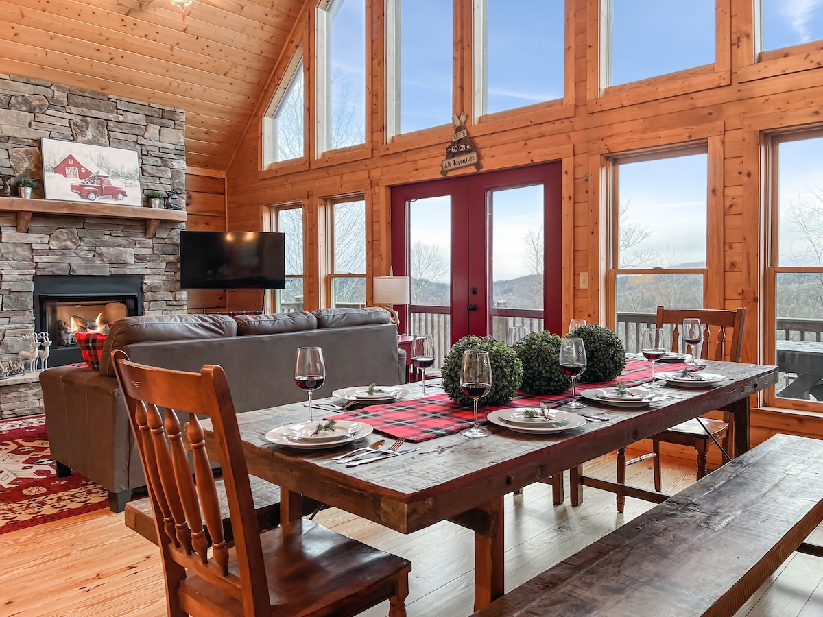 An open-concept living space is highlighted by floor-to-ceiling windows framing mountain views. A large wooden dining table, set for six, is adorned with elegant dishware. A cozy sofa is positioned near a stone fireplace, offering a welcoming ambiance for gatherings.