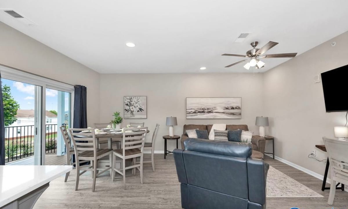 The open living space features a comfortable seating area with a sectional sofa and a lounge chair. A dining table is positioned nearby, accommodating several seats. Natural light fills the area through sliding doors that lead to a balcony, creating a bright atmosphere.