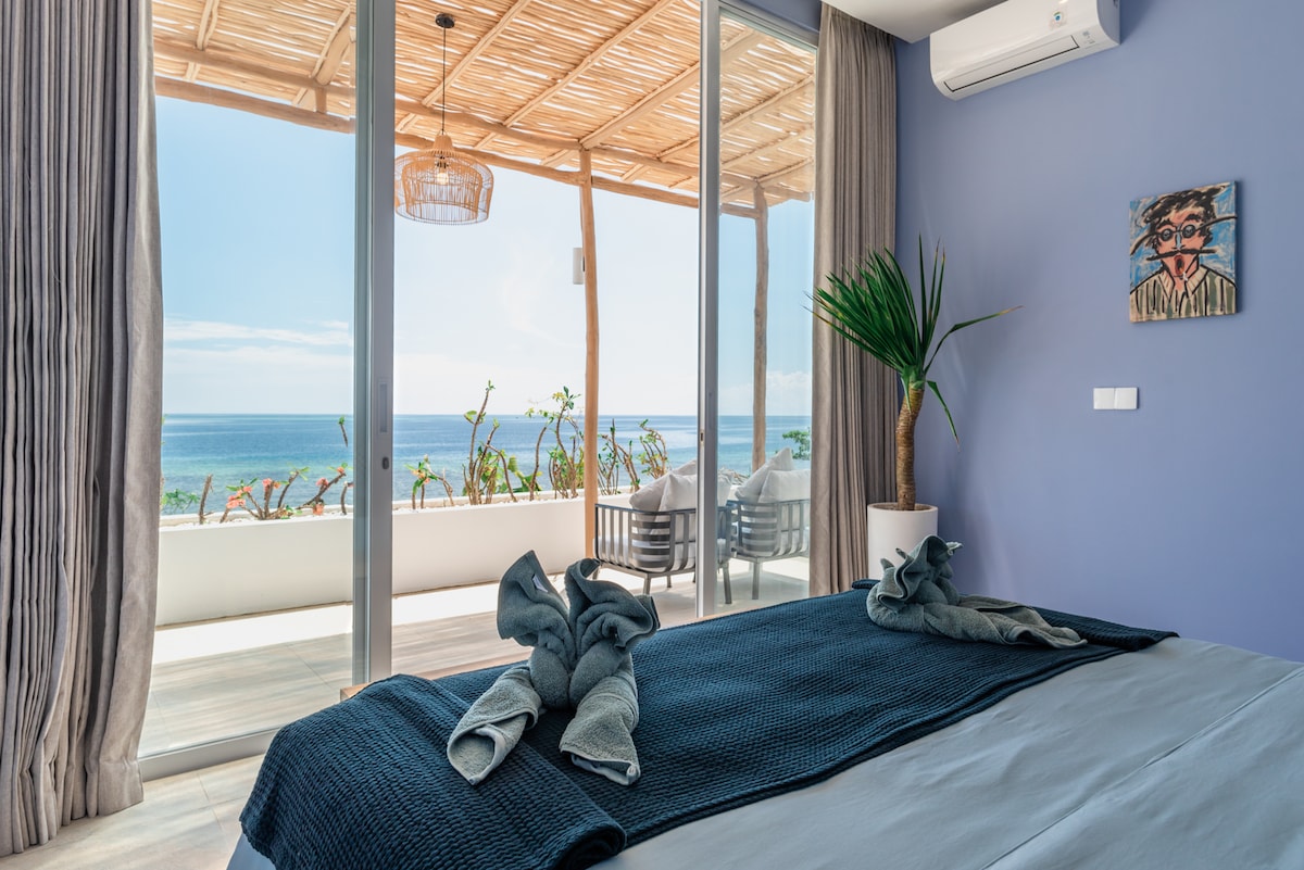 A light-filled bedroom features a spacious bed with rolled towels at the foot. Floor-to-ceiling glass doors open to a private balcony overlooking the ocean. Natural elements, such as a potted plant and wicker lighting, complement the tranquil blue walls.