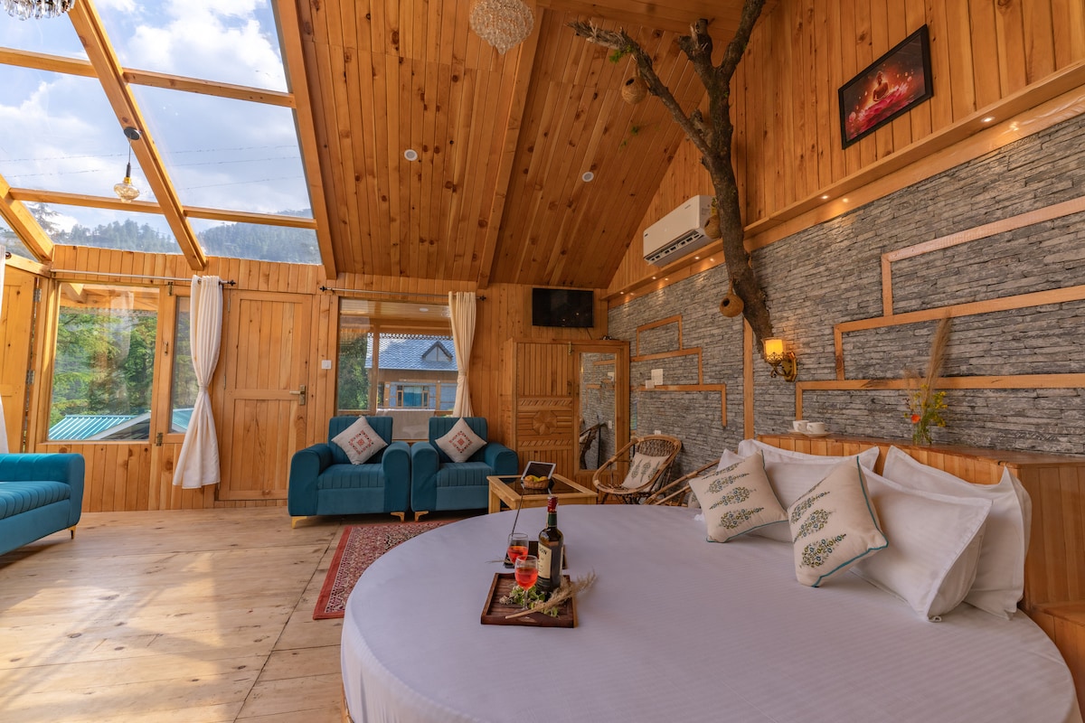 A spacious room features wooden walls and a high ceiling, creating a warm ambiance. A large round bed is positioned in the center, surrounded by comfortable seating areas with blue sofas. Natural light filters through large windows, enhancing the overall inviting space.