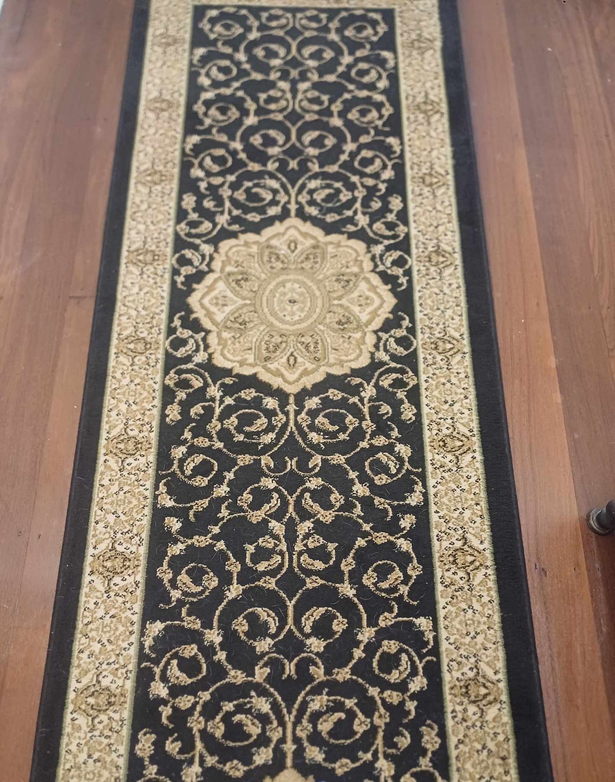 A long, decorative runner rug in black and gold features intricate floral and vine patterns. The rug is positioned on polished wooden flooring, enhancing the elegant ambiance of the space.
