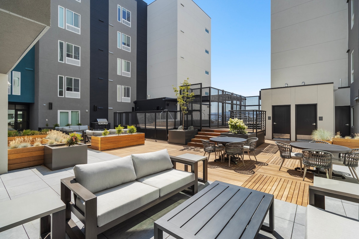 An outdoor patio space is presented, featuring wooden decking with various seating arrangements, including chairs and tables. Planters with greenery add natural elements, while a clear sky enhances the modern building's sleek architecture in the background.