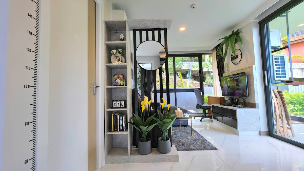 A modern living area is seen with large windows allowing natural light. Decorative elements include a full-length mirror, a small bookshelf filled with books, and vibrant yellow flowers in planters. A cozy seating area complements a sleek television unit against a contemporary backdrop.
