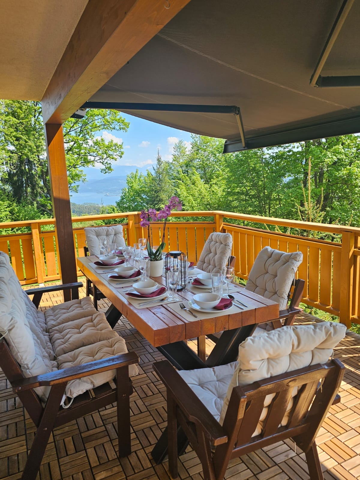 An outdoor dining area is showcased, featuring a wooden table surrounded by six cushioned chairs. A view of lush greenery and distant mountains is visible through the railing. The table is set with plates and glasses, inviting casual meals in a serene environment.