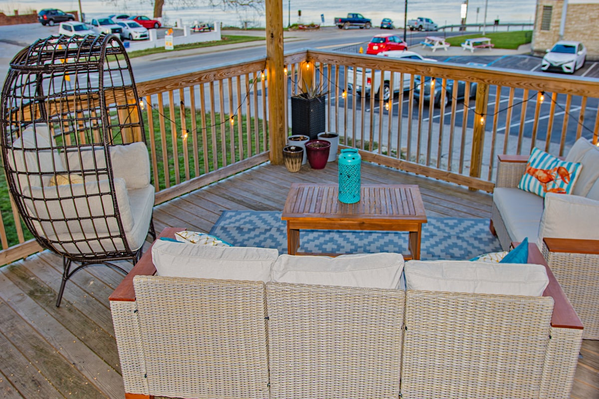 A private deck is shown with cozy seating arrangements, featuring a light-colored wicker sofa and an inviting hanging chair. A wooden coffee table, accented by a decorative blue vase, is positioned at the center. String lights add a soft glow, enhancing the overall ambiance.