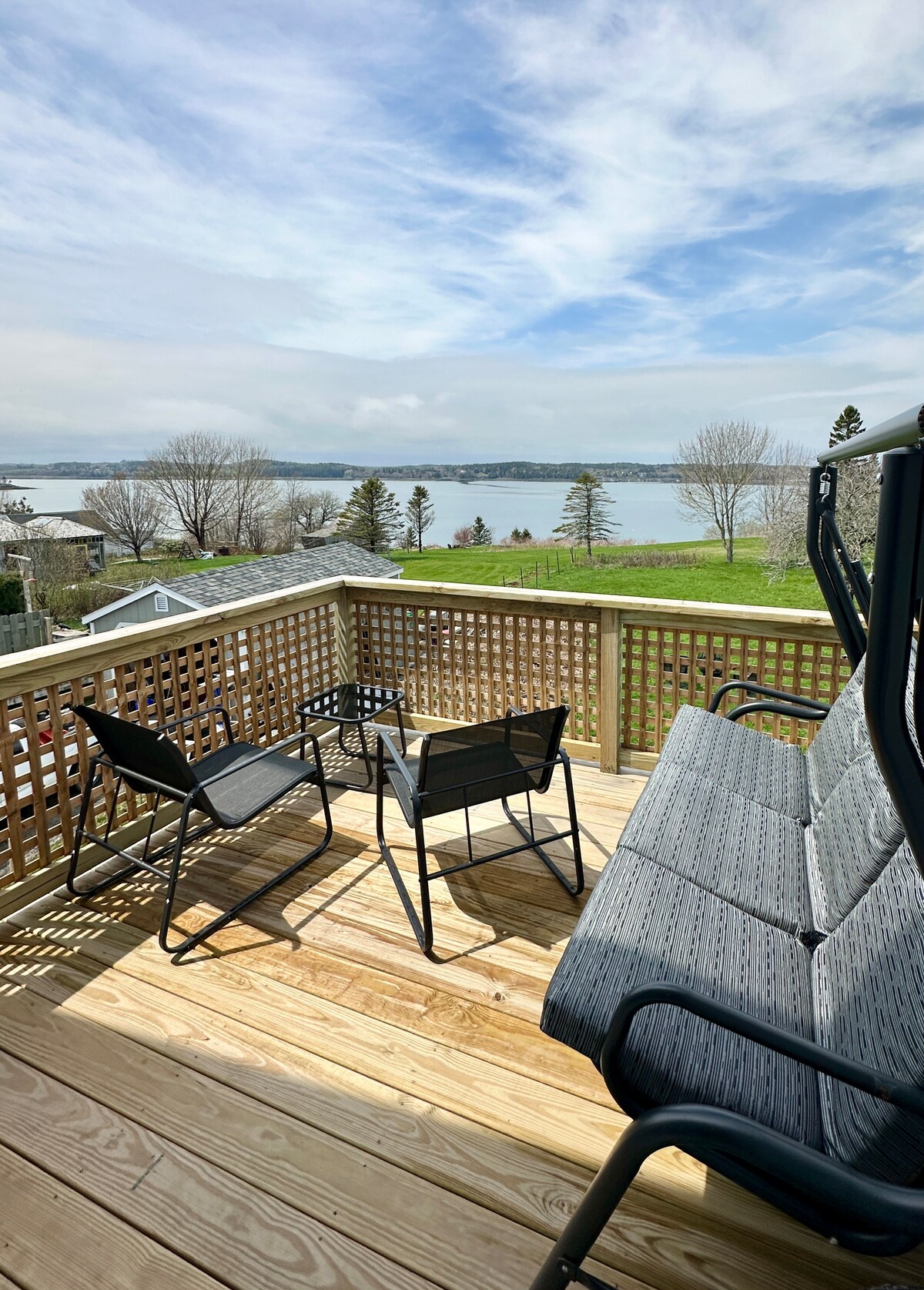 A wooden deck is presented with seating options, including a glider and two chairs, overlooking Johnsons Bay. The area is bathed in natural light, and grassy land extends beyond the deck, with water and trees visible in the distance.