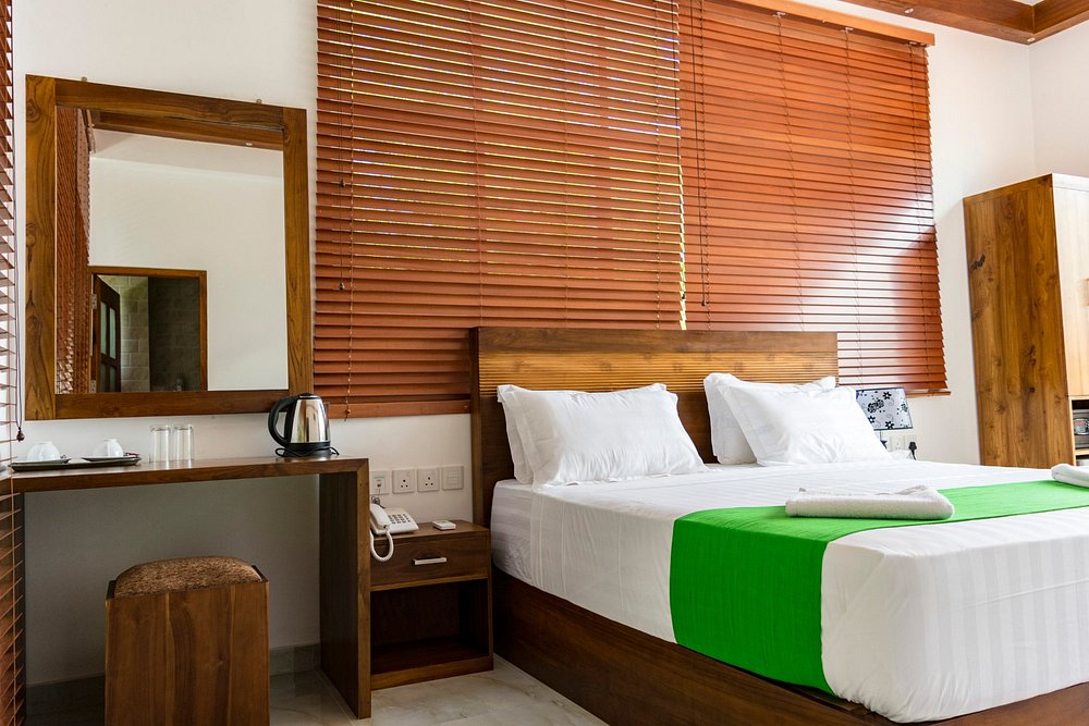 A bright room features a spacious bed with crisp white linens and a green runner. Natural wood furniture includes a desk and a mirror, while horizontal blinds provide control over light entry. A kettle is placed on the desk, complementing the minimalist decor.