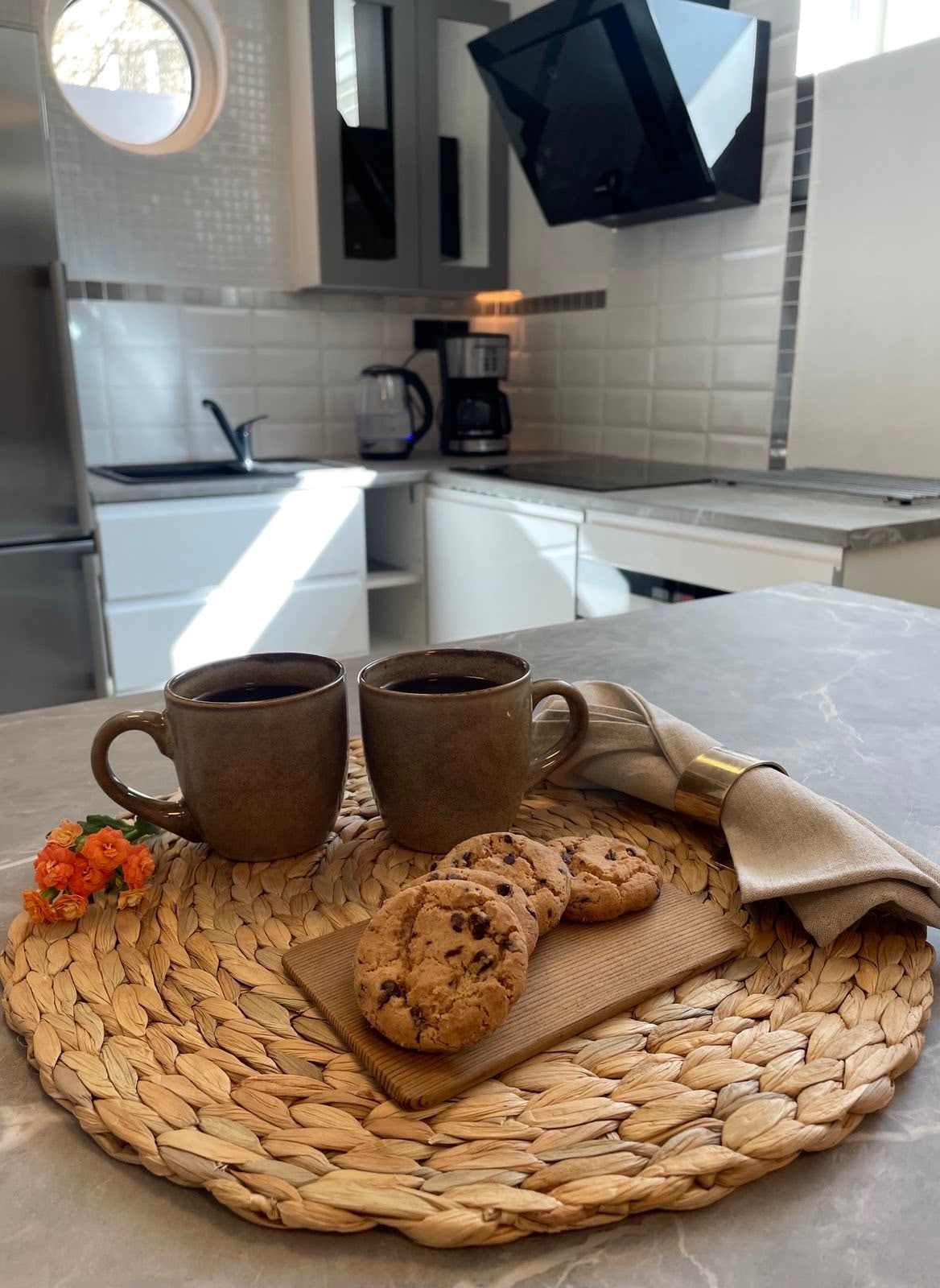 A kitchen countertop displays two ceramic mugs filled with coffee beside freshly baked cookies on a wooden serving board. A woven placemat holds the cookies, adding a natural element to the scene. Soft light filters through nearby windows, illuminating the modern kitchen space.