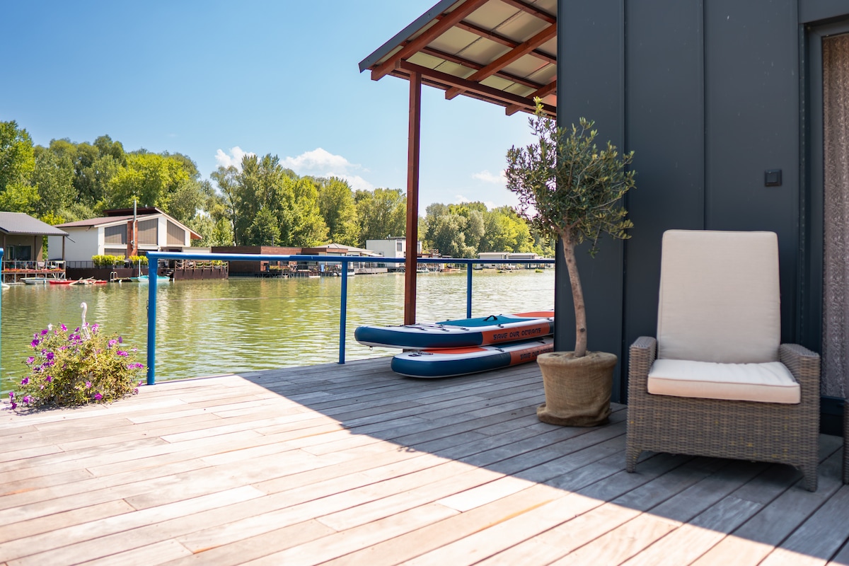 A spacious sun terrace overlooks the serene waters of the Danube bay. Two paddleboards are positioned nearby, while a comfortable chair sits next to potted greenery. The scene features wooden decking and a clear sky, inviting outdoor relaxation and exploration.