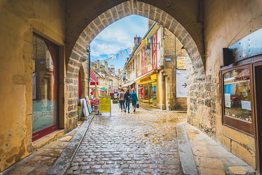 A charming cobblestone passageway lined with historic buildings leads to a vibrant street filled with colorful awnings and decorations. Several people can be seen strolling by, enjoying the lively atmosphere. The sky above is partly cloudy, allowing natural light to illuminate the scene.