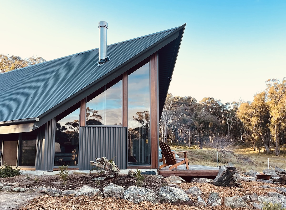 The exterior of a contemporary cabin featuring a sloped roof and large glass windows. A wooden chair sits on the deck beside natural landscaping of rocks and mulch, with a backdrop of trees and open land.