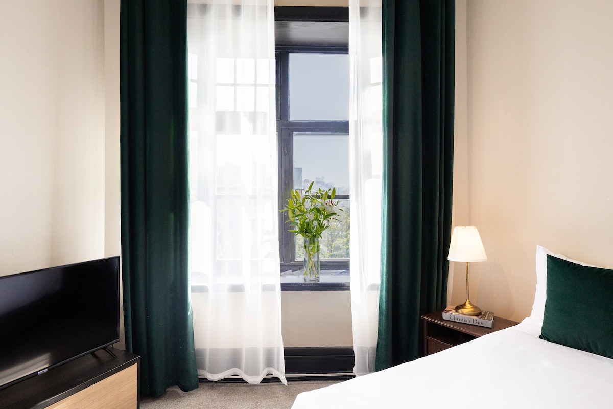 A cozy room features a spacious bed adorned with quality linens. Natural light is filtered through sheer and dark curtains, illuminating a small table with a vase of fresh flowers. A flat-screen TV sits on a low wooden dresser, enhancing the room's functionality.