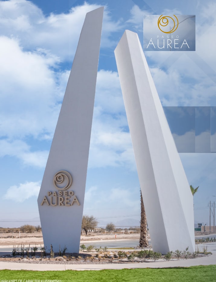The entrance to Paseo Aurea is marked by two tall, modern white structures that rise prominently against a clear sky. A circular logo is displayed at the top, and the name 'Paseo Aurea' is featured at the base, surrounded by neatly landscaped greenery.