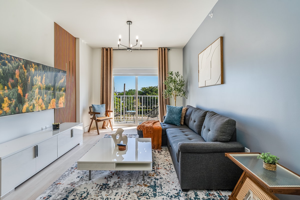 A spacious living room features a comfortable sectional sofa facing a large television mounted on the wall. Natural light enters through glass doors leading to an outdoor balcony, which is complemented by modern decor, elegant curtains, and a stylish coffee table.