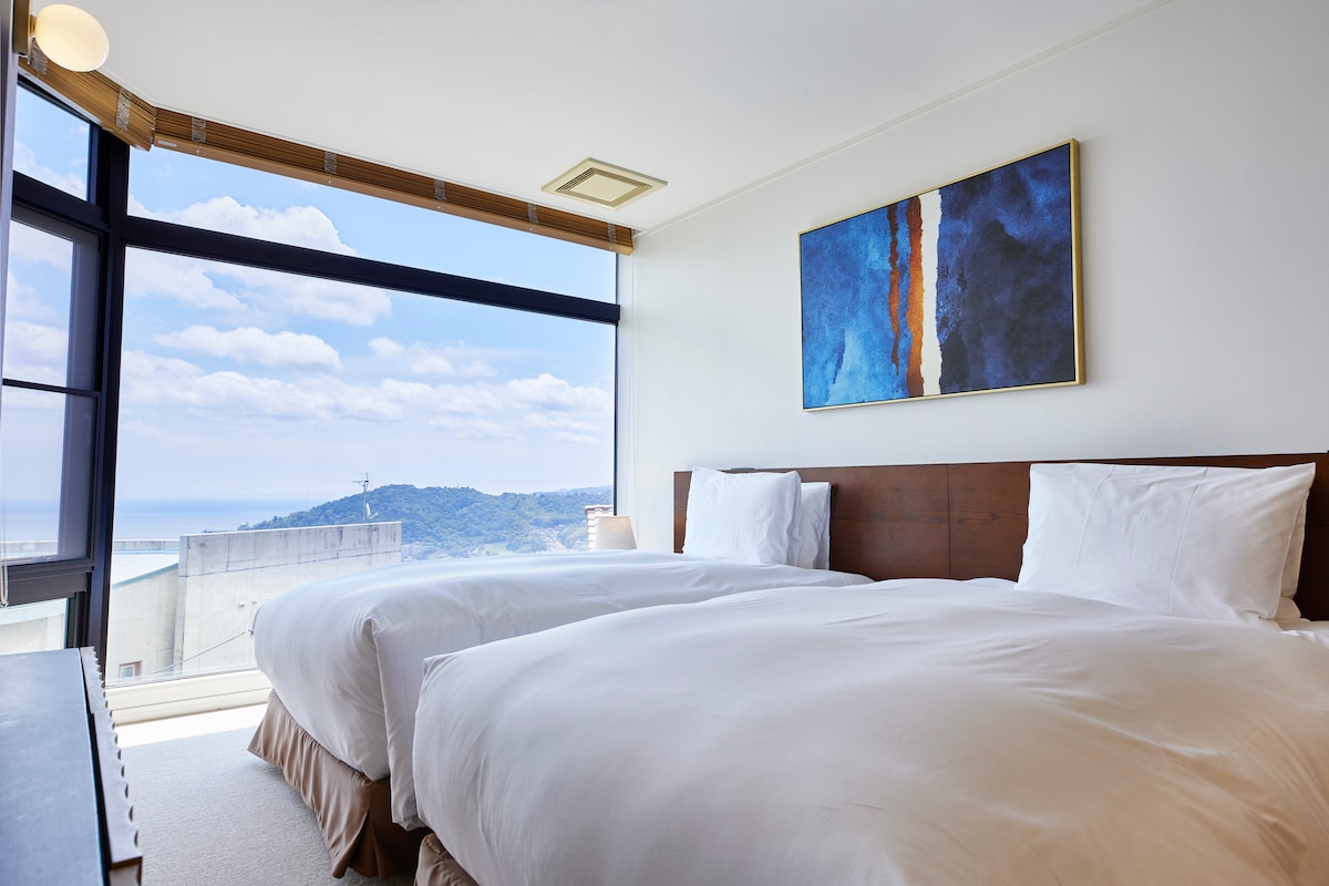 A light-filled bedroom is presented with two spacious beds arranged side by side. A large window offers a panoramic view of the surrounding hills and ocean, while an abstract art piece adds a touch of color to the neutral walls.