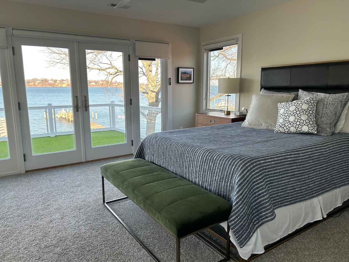 A sunlit bedroom features a spacious bed, dressed in neutral tones with decorative pillows. A green bench is positioned at the foot. Double doors open to a scenic view of the waterfront, while soft carpeting adds warmth to the space.