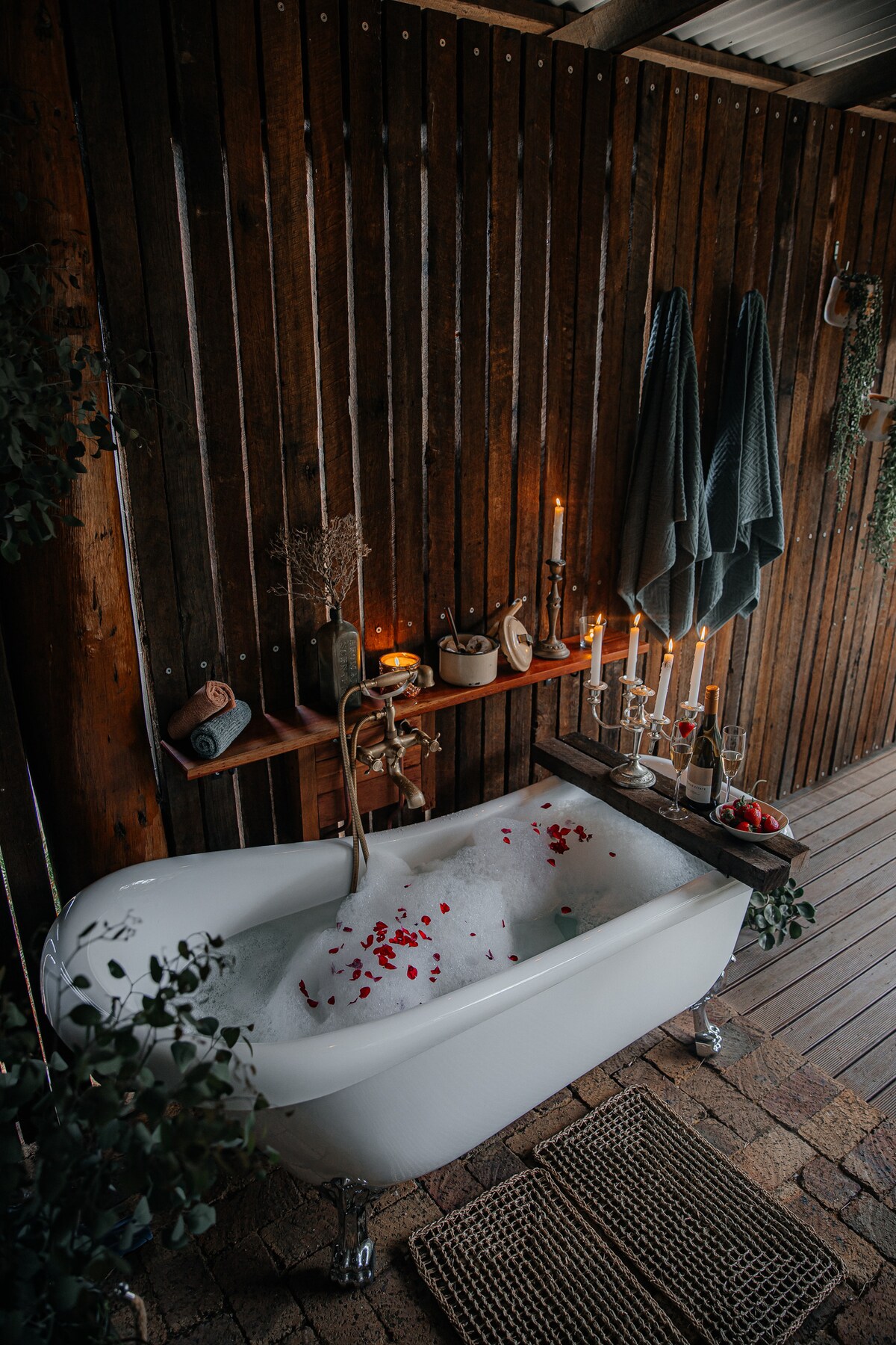 An outdoor claw foot bathtub is displayed against a wooden wall, surrounded by lush greenery. The tub is filled with bubbles and adorned with rose petals. Soft candlelight illuminates nearby shelves decorated with bath essentials and a romantic touch of fruit.
