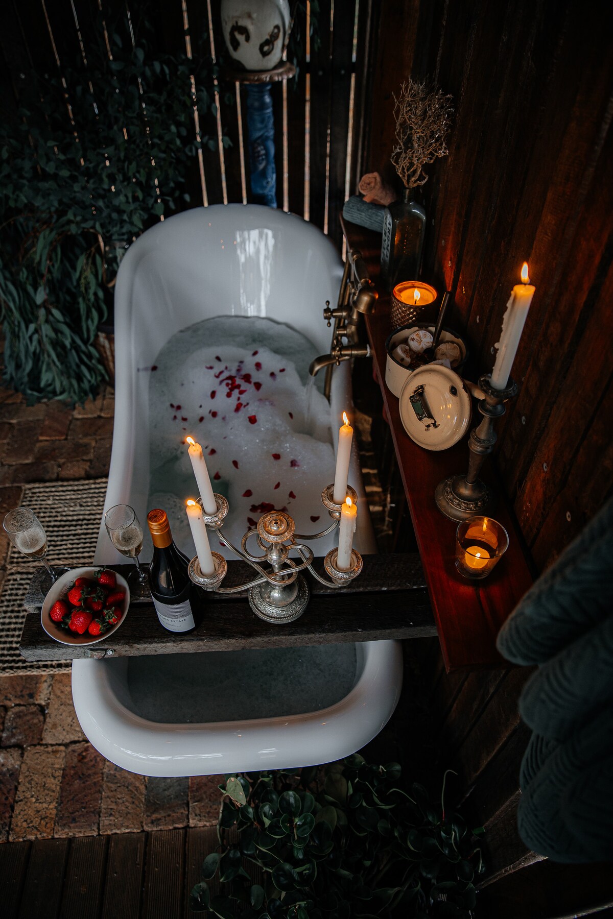 An outdoor clawfoot bathtub is partially filled with bubbles and rose petals, surrounded by greenery. Candles of varying heights cast a warm glow, while a small serving tray with strawberries and a bottle of sparkling wine is placed nearby for an inviting touch.