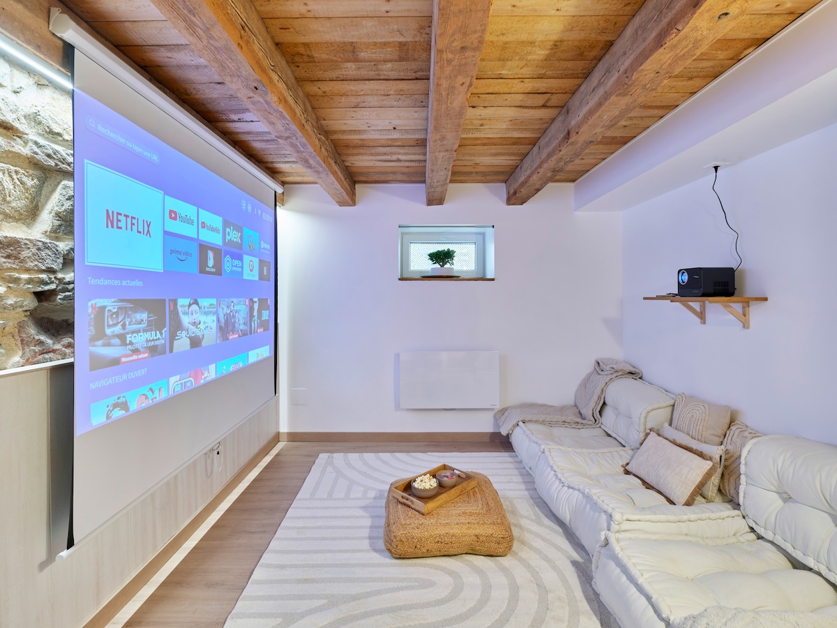 A cozy entertainment space is presented, featuring a large projection screen displaying streaming options. A comfortable sectional sofa with soft cushions is arranged around a stylish woven coffee table. Warm wooden beams add a rustic touch to the modern decor, enhancing the welcoming atmosphere.