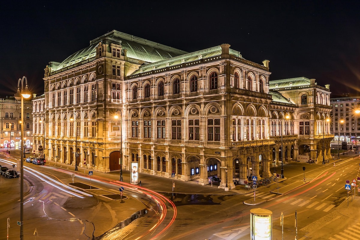 An illuminated historical building stands prominent at night, showcasing intricate architectural details and a dome roof. Surrounding streets are marked by flowing light trails from passing vehicles, adding a sense of movement to the tranquil scene. The atmosphere reflects Vienna's cultural elegance.