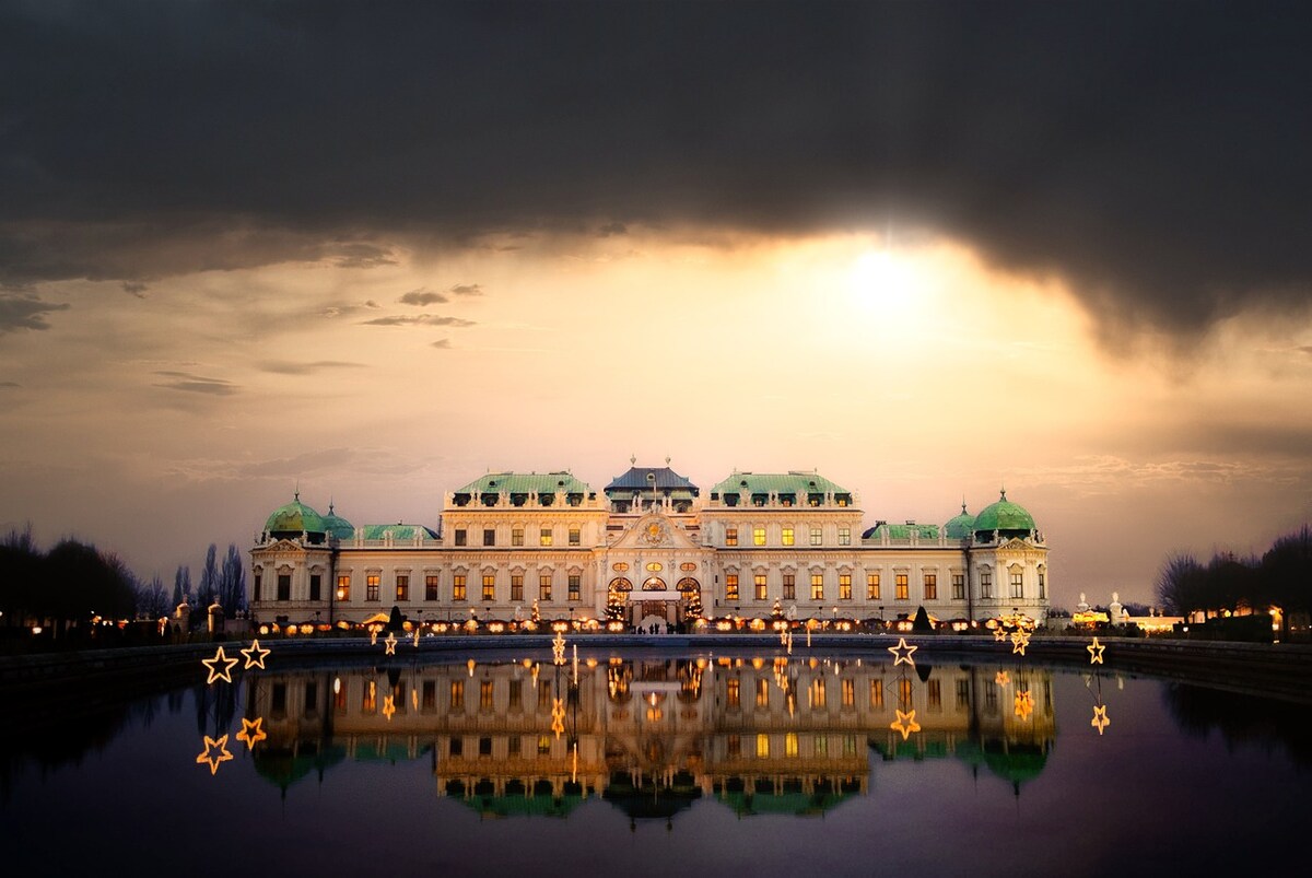 A grand historic building is reflected in a serene water feature, with ornamental lights positioned around the perimeter. The structure is characterized by a symmetrical façade, green domes, and detailed architectural elements, framed by a twilight sky that hints at an impending sunset.