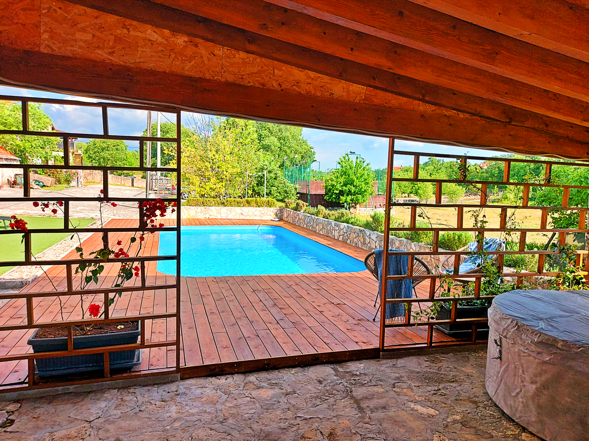 A spacious outdoor area features a clear blue pool surrounded by a wooden deck. Natural light fills the space through a decorative wall of glass blocks. Potted plants with flowers add greenery, while a hot tub is partially visible on the right side.