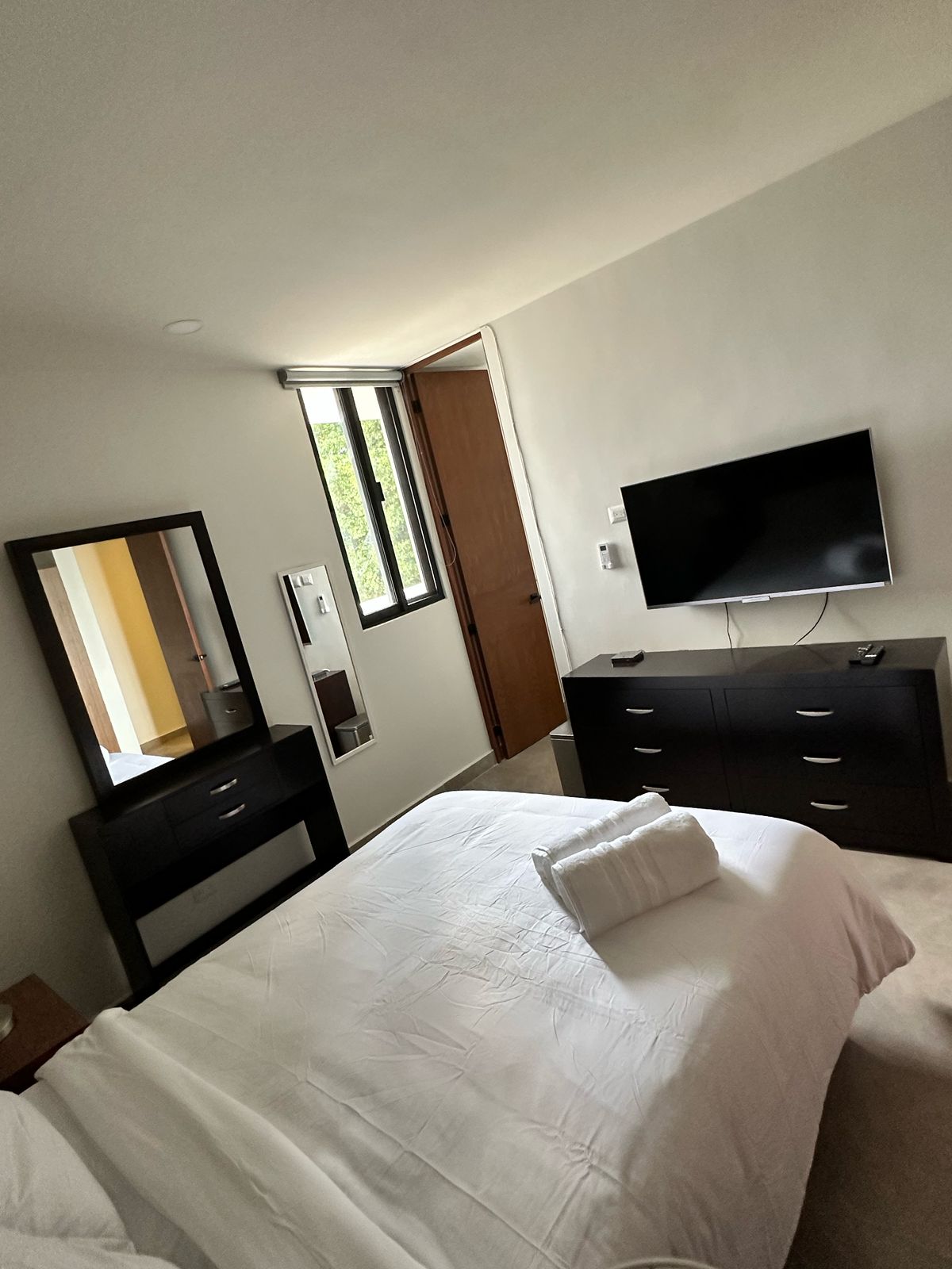 The bedroom features a spacious bed dressed in white linens, complemented by a white decorative pillow. A large flat-screen TV is mounted on the wall opposite the bed. A contemporary dresser and a mirror with a dark frame occupy one side of the room, while natural light enters through a window.