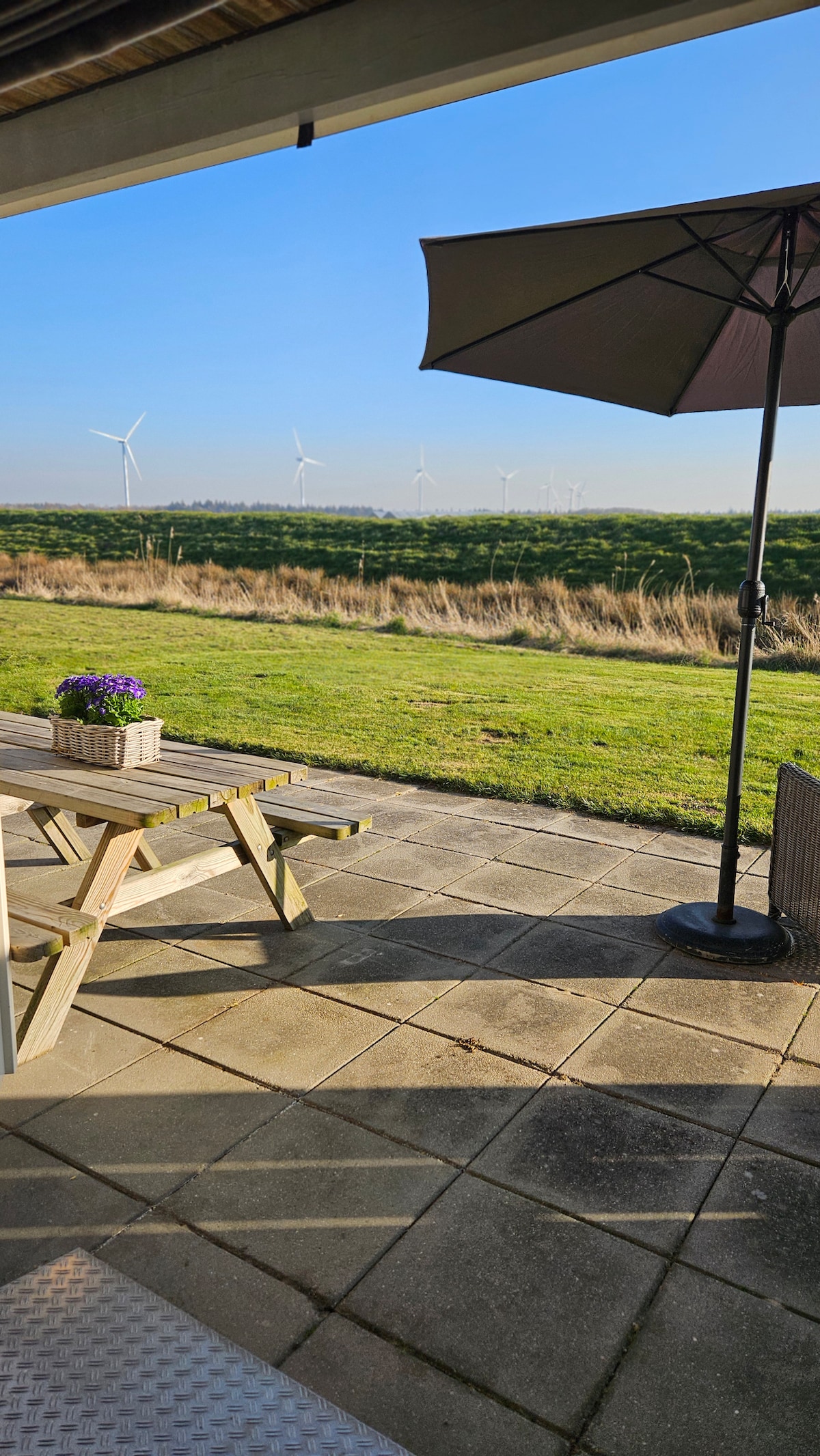 An outdoor terrace is presented with a wooden picnic table and a large parasol providing shade. A planter with flowers sits atop the table, while a grassy area extends toward a view of wind turbines in the distance under a clear blue sky.