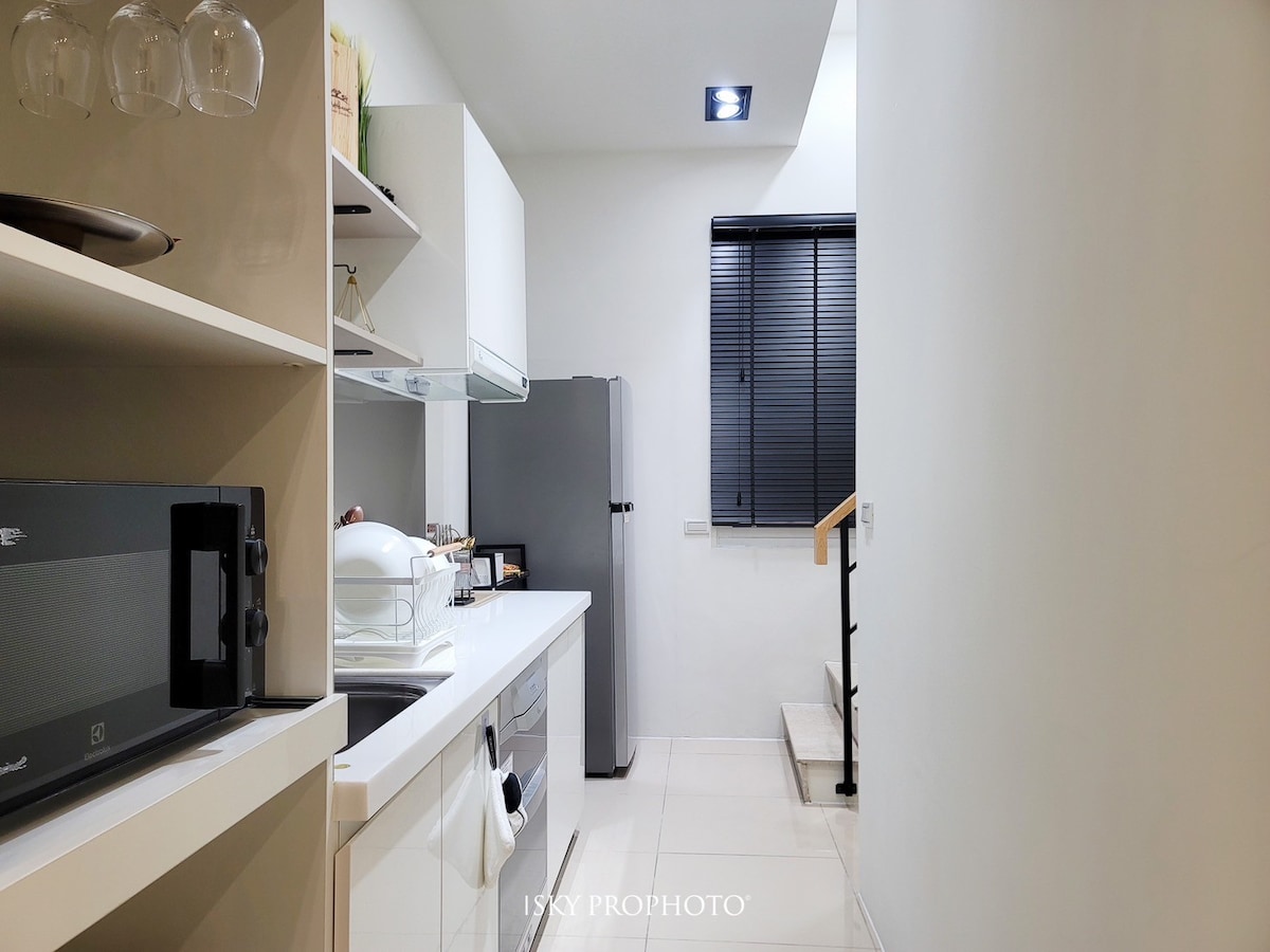 A modern kitchen area is depicted, featuring white cabinetry and a countertop. Essential appliances, including a microwave and a refrigerator, are visible. A window with blinds allows natural light into the space. Stairs leading to another level can be seen at the end of the hallway.