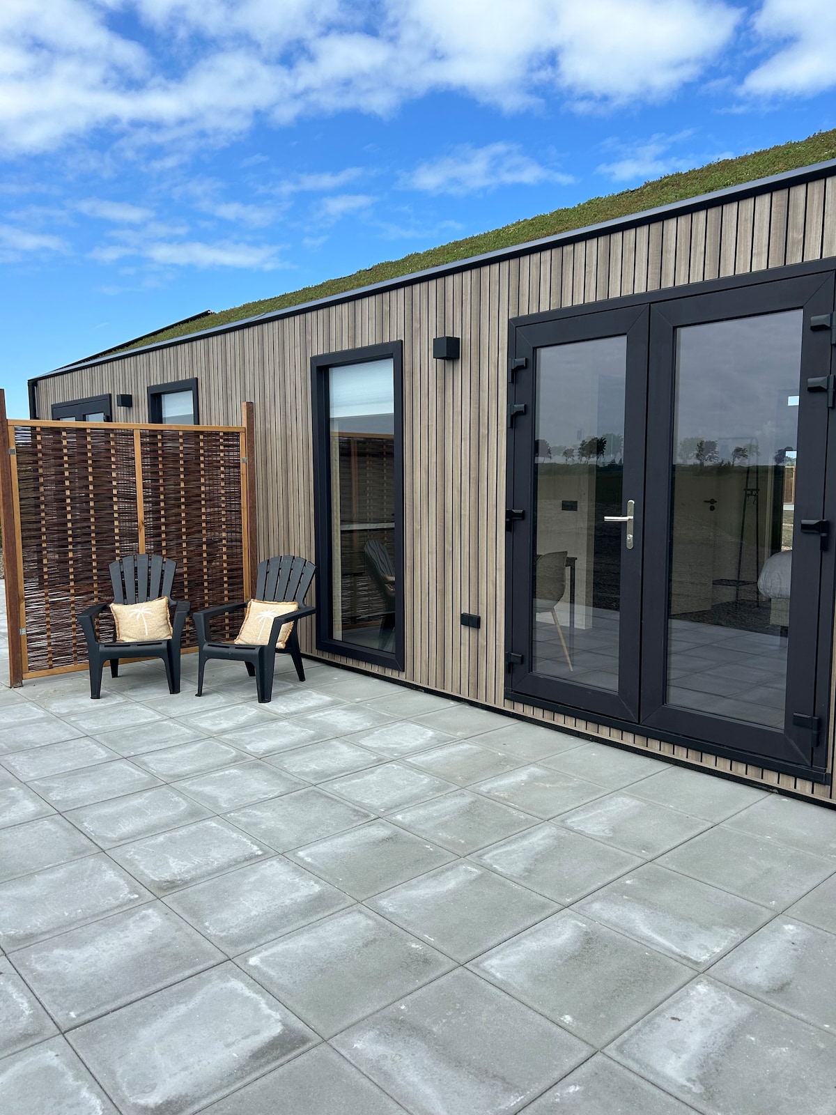 A compact outdoor patio area features two black chairs positioned beside a wooden privacy screen. The facade of the lodge, characterized by vertical wooden slats and large glass doors, is visible against a backdrop of a clear blue sky.