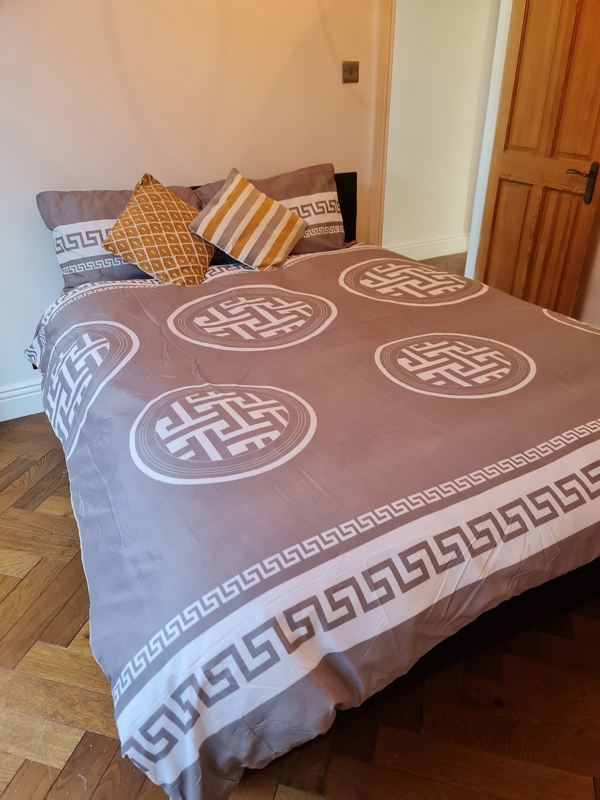 A spacious bed is adorned with a patterned duvet featuring circular designs and a Greek key border. Two accent pillows, one with stripes and the other in a solid color, are arranged at the head of the bed. Warm wooden tones are visible in the door and flooring.