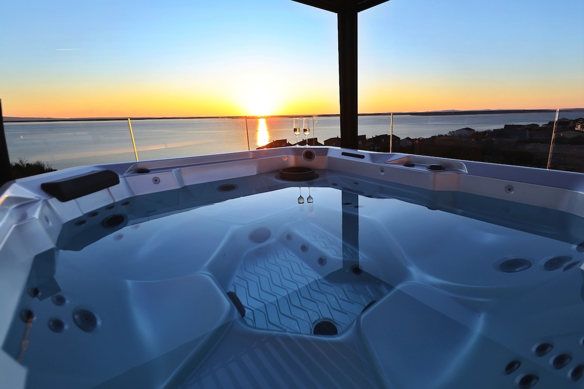 A private jacuzzi is situated on a terrace, offering a view of the setting sun over the water. The calm surface of the water reflects the warm tones of the sunset, while nearby glass railings provide an unobstructed panoramic view of the surrounding landscape.
