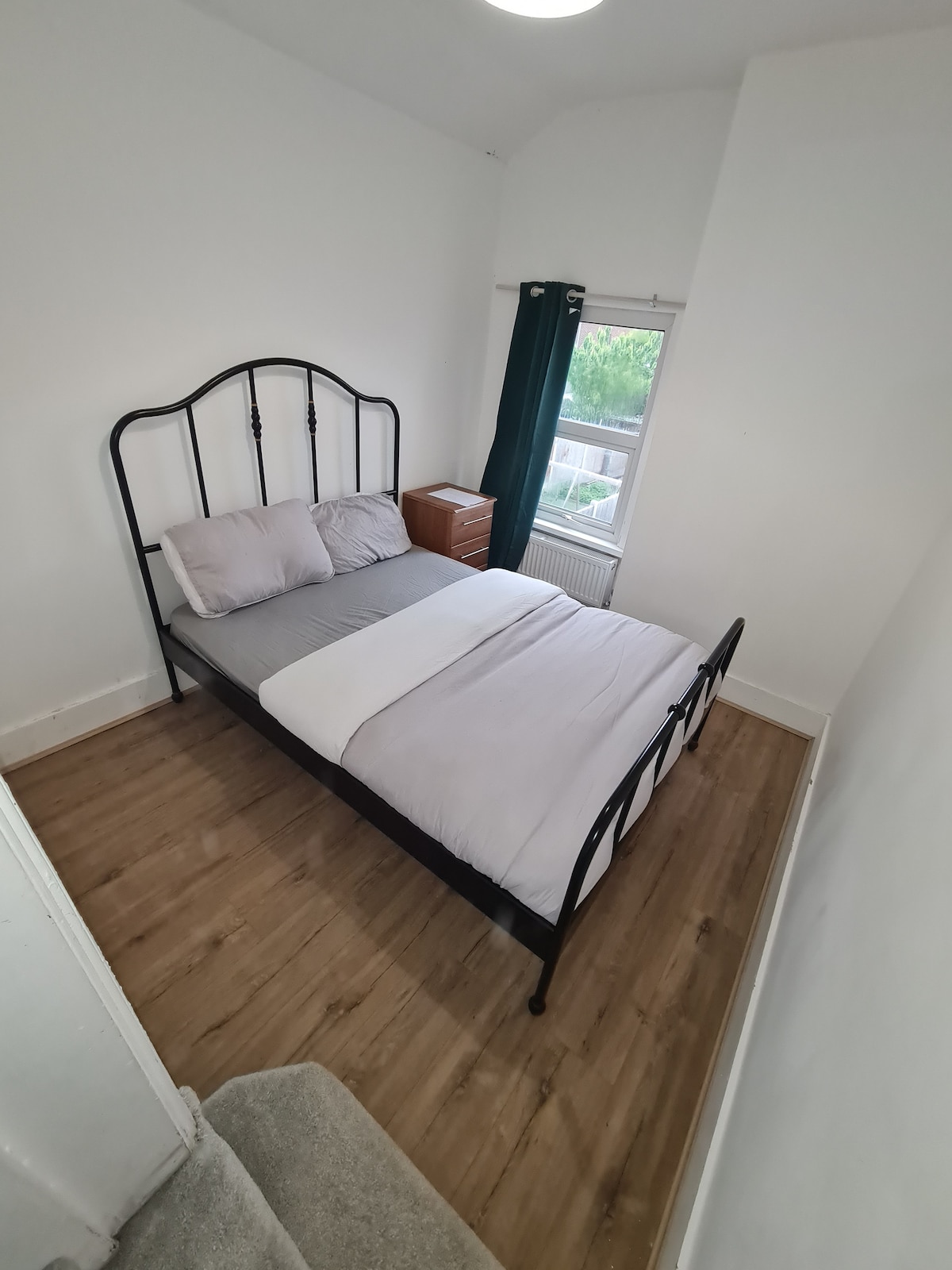 A cozy sleeping area features a black metal bed frame with a plush light grey comforter. Nearby, a wooden bedside table supports a small lamp. Natural light enters through a window adorned with dark green curtains, adding to the room's inviting ambiance.