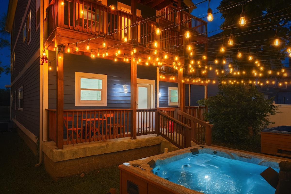 The exterior of the home is illuminated by warm string lights, creating a cozy ambiance at night. A private hot tub is situated on the ground level, surrounded by greenery, while two upper balconies provide additional outdoor seating options.