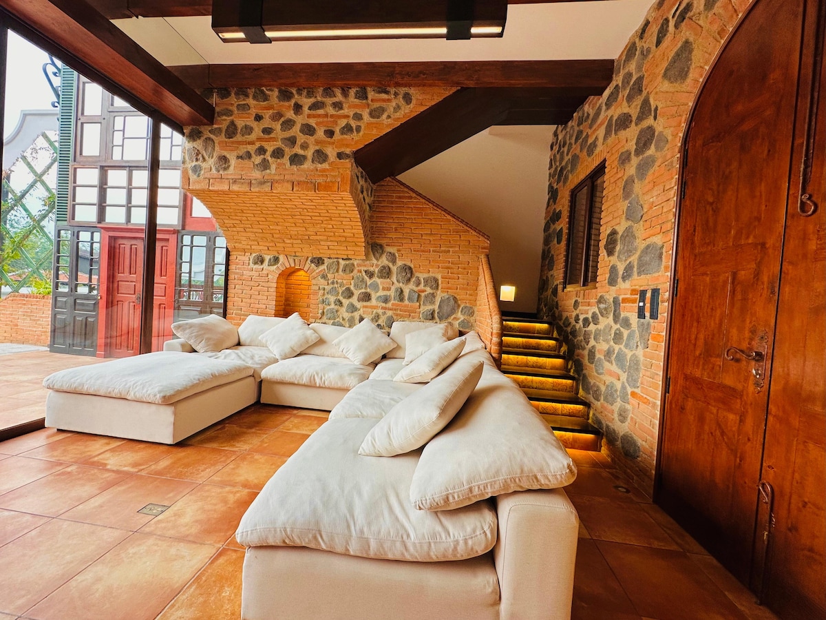 An inviting seating area features a series of plush, neutral-toned sofas arranged for comfort. The textured stone walls and wooden beams create a rustic charm, while a staircase leads to a well-lit space, enhancing the overall warmth of the room.