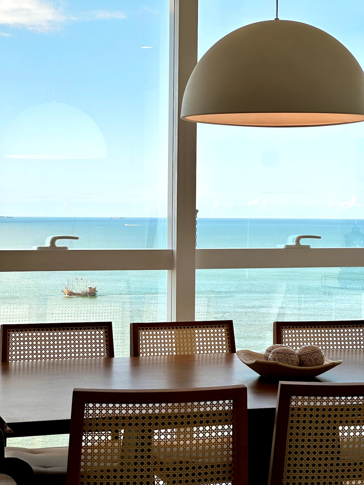 A dining area is presented with a wooden table surrounded by woven chairs. A large light fixture hangs overhead, illuminating the space. Through expansive windows, a panoramic view of the ocean is visible, with distant boats enhancing the coastal atmosphere.