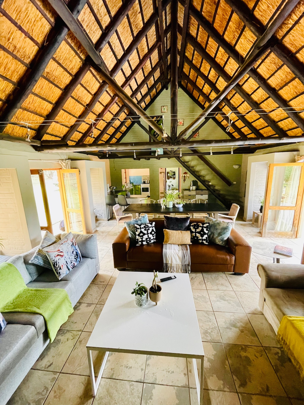 A spacious living area is shown with high, thatched ceilings and exposed wooden beams. Comfortable seating includes a mix of colorful cushions on sofas. A central coffee table is placed on tiled flooring. Bright sunlight streams through glass doors that open to the outdoor area.