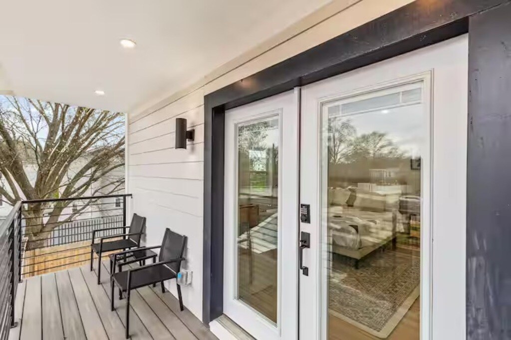 A private balcony is showcased with two black chairs facing outward. Glass sliding doors provide a view into a well-lit living area featuring a beige sofa and decorative rug. The outdoor space features light wood flooring and offers a serene setting for relaxation.