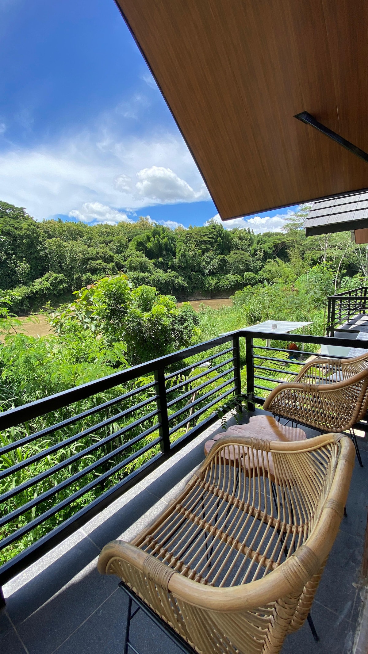 A balcony area features two rattan chairs facing a lush green landscape. The natural scenery includes a river bordered by dense vegetation under a partly cloudy sky. Sunlight bathes the space, enhancing the outdoor seating experience.