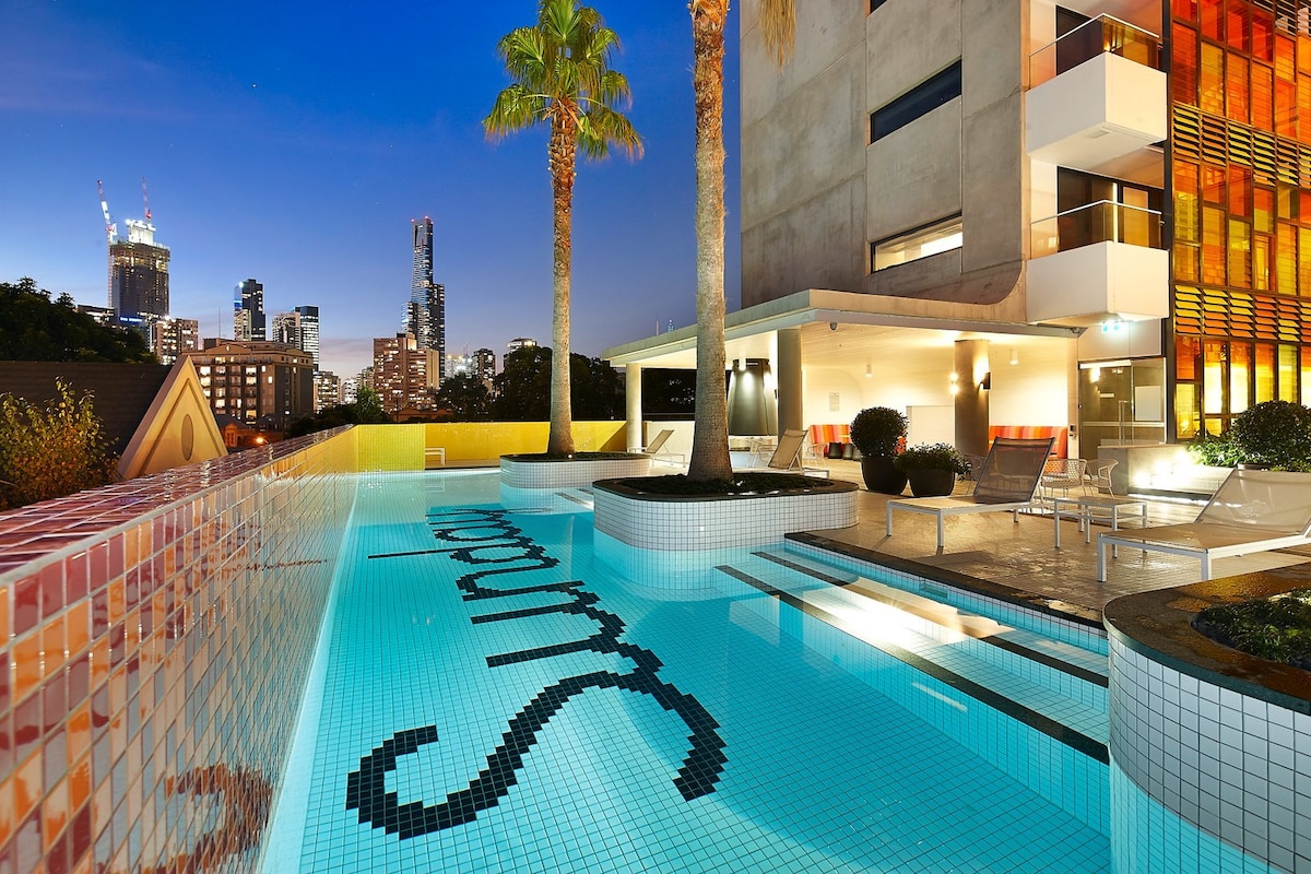 A tranquil swimming pool is presented, featuring vibrant mosaic tiles that spell 'Sunday.' Surrounding palm trees and modern architecture enhance the outdoor space, while the evening city skyline provides a stunning backdrop, illuminated by city lights.