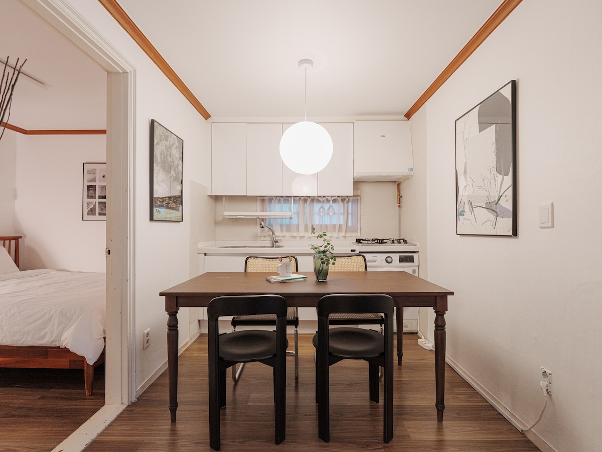 A dining area features a dark wooden table accompanied by four black chairs. A pendant light hangs above, illuminating the space. In the background, a modern kitchen setup with white cabinetry and a stove is visible, along with artwork displayed on the wall.