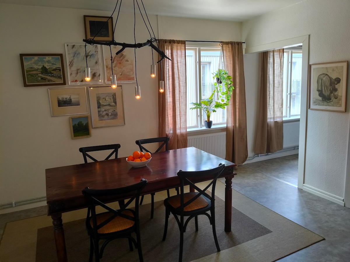 A dining area is present with a wooden table centrally located, surrounded by four chairs. An arrangement of fruit sits in the middle of the table. Walls are adorned with framed artwork, and large windows are dressed with light brown curtains, allowing ample natural light.