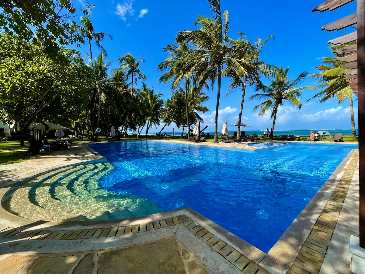A large swimming pool reflects the clear blue sky and is surrounded by lush palm trees. The area features a gentle slope leading into the pool, with lounge chairs positioned under shaded umbrellas nearby. The ocean is visible in the background, enhancing the tranquil setting.