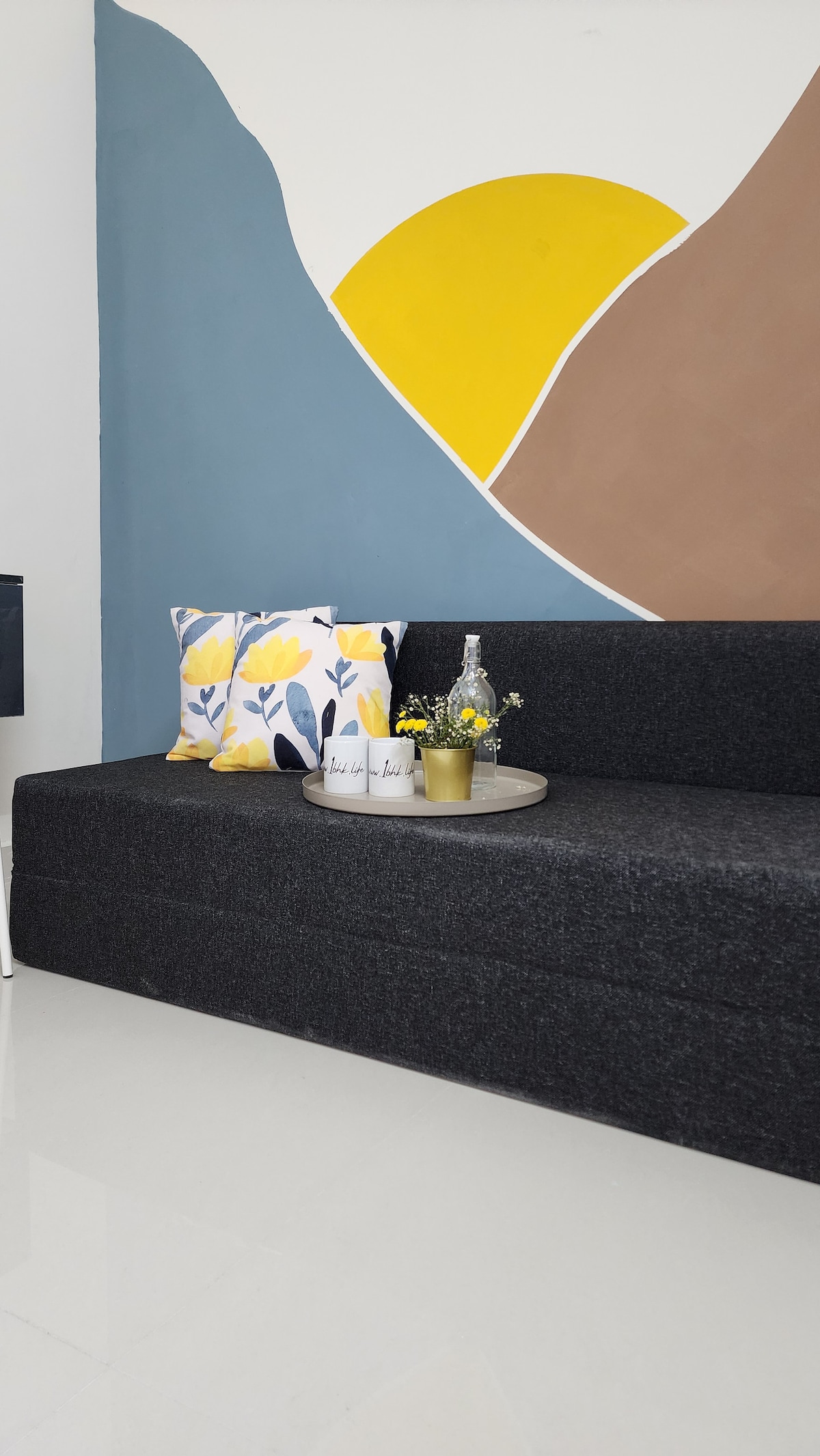 A modern living area is depicted with a dark gray sofa positioned against a stylized wall featuring abstract shapes in yellow and brown. A round tray holds decorative items and a small vase with flowers. Colorful throw pillows add a pop of floral design.