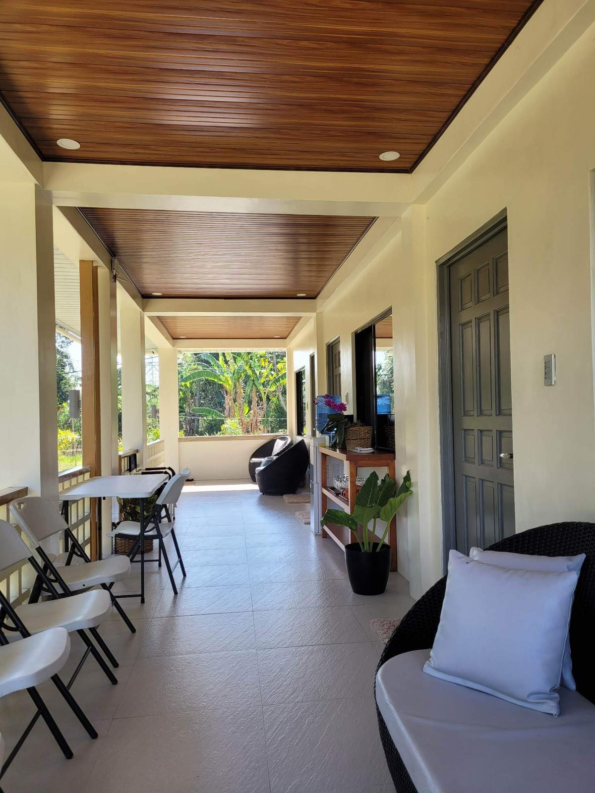 A spacious porch area features a smooth tile floor and natural wood ceiling. Light-colored chairs are arranged along one side, while comfortable seating invites relaxation. Large windows allow views of the landscape, enhancing the connection to nature.