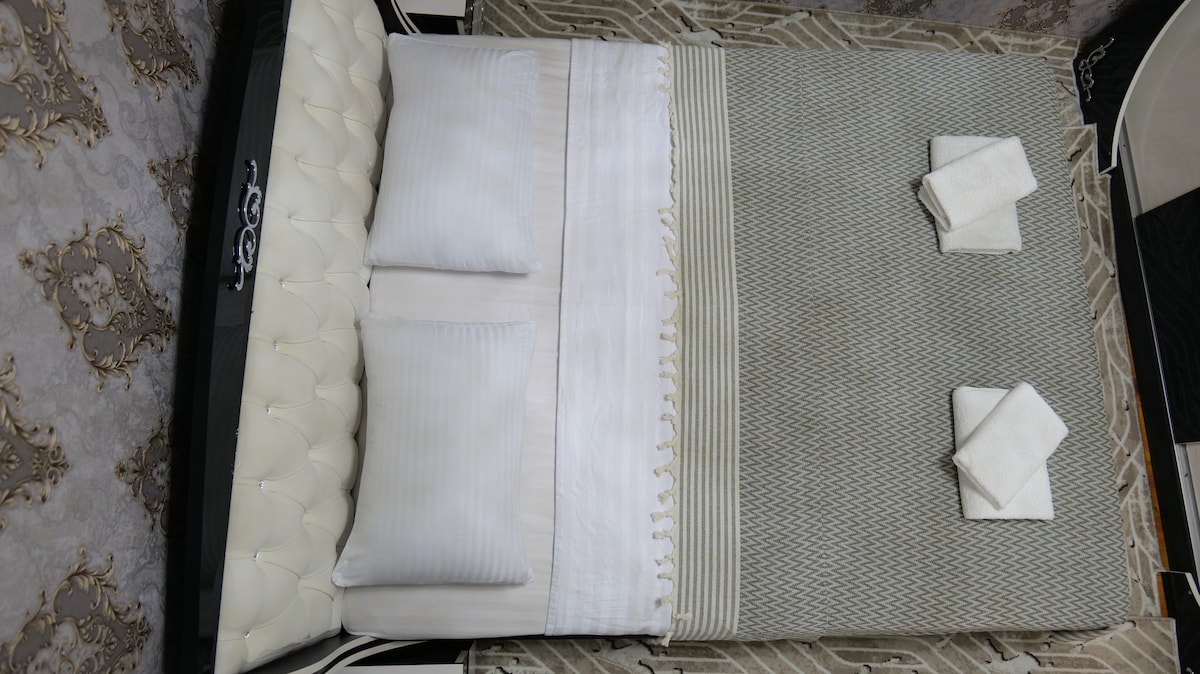 A spacious double bed is presented with neatly arranged pillows and towels. The elegant bedding features a subtle pattern, complemented by a tufted headboard. Soft, neutral tones create a calming atmosphere, while the surrounding decor adds a touch of sophistication.