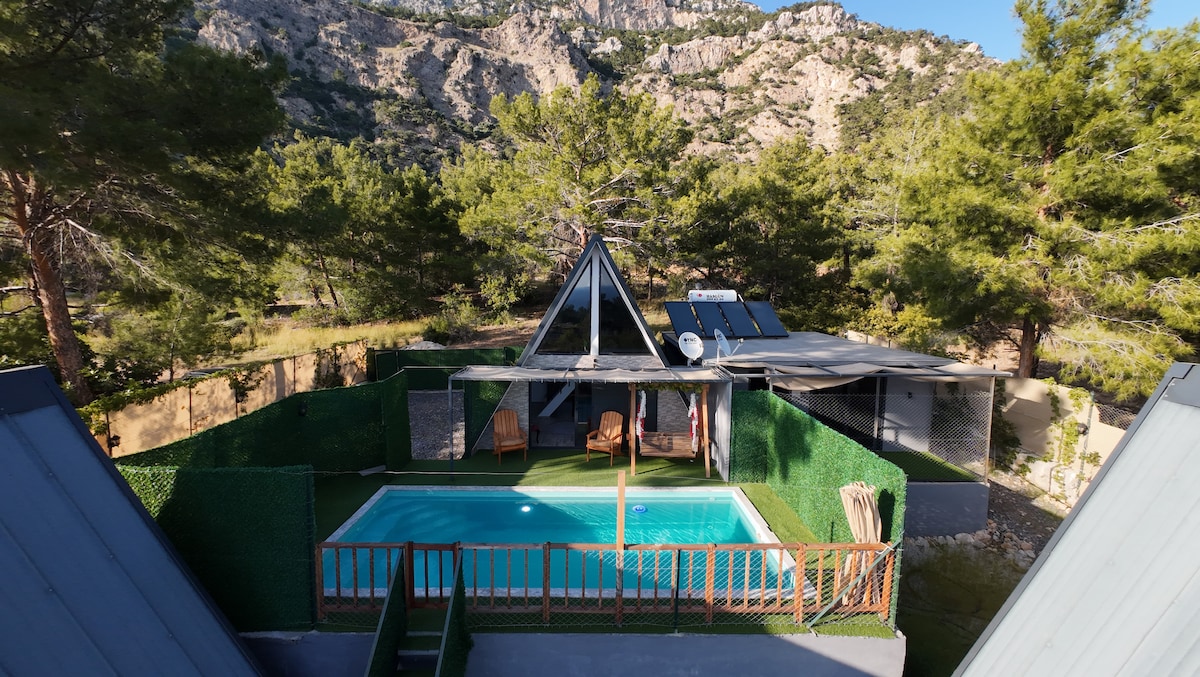 An outdoor area features a swimming pool surrounded by lush greenery. Two wooden lounge chairs are positioned beside the pool, offering a place to relax. The backdrop consists of mountains, and a triangular-roofed structure is visible in the distance.