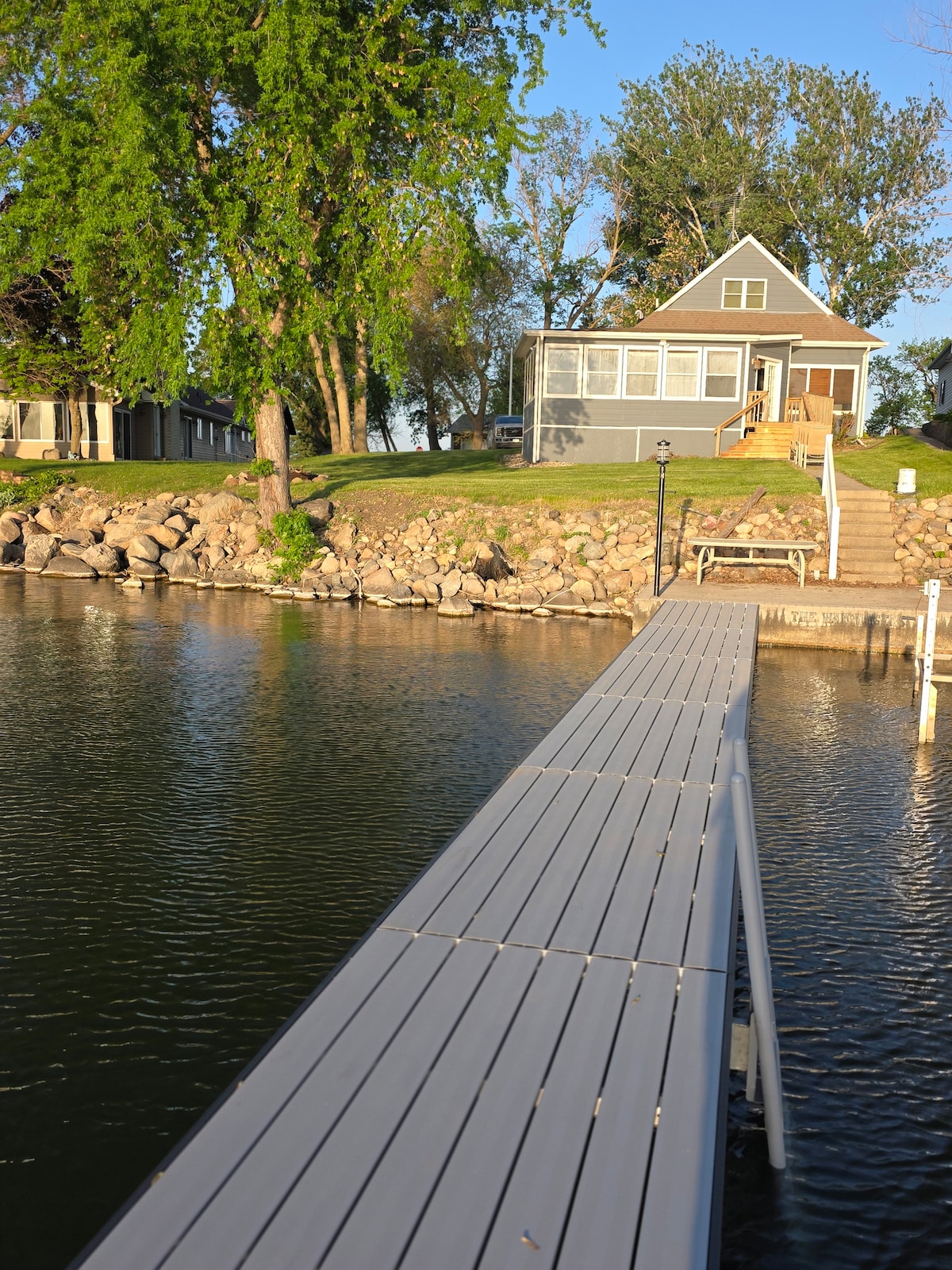A wooden dock extends over the water, leading to a gray two-story house with a welcoming sunroom. Lush greenery and large rocks line the shoreline, while the calm lake reflects the tranquil surroundings.