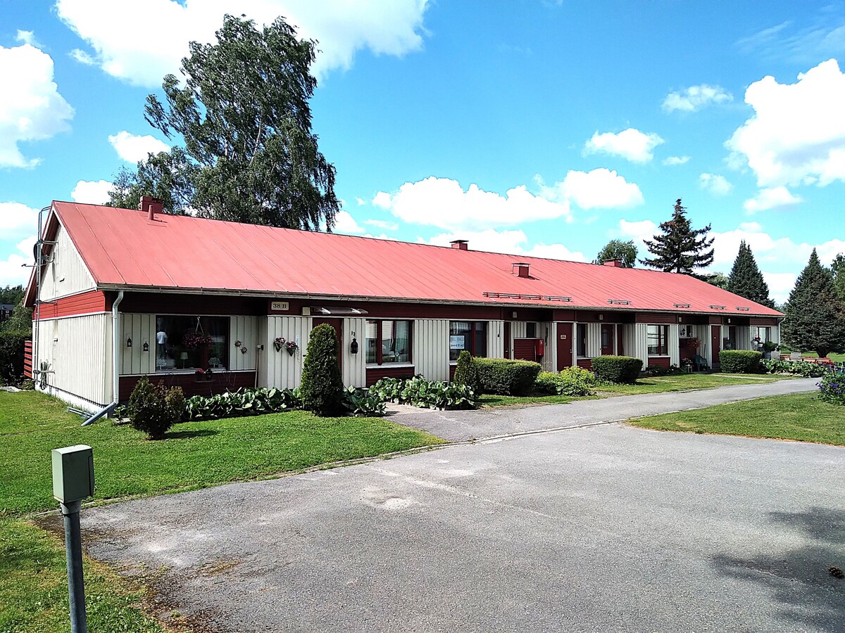 A row of charming townhouses is depicted, featuring a red roof and light-colored siding. Lush greenery lines the pathway, which leads to each unit. The sky is bright with scattered clouds, creating a welcoming atmosphere for residents and visitors.