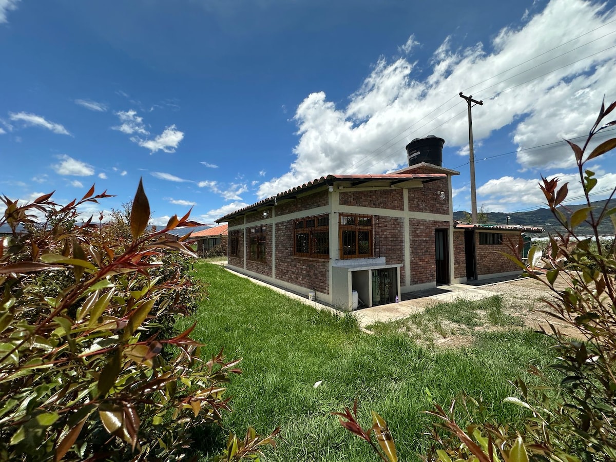 A single-story cabin is framed by lush green grass and shrubbery. Large windows allow natural light to fill the interior, while a stone chimney extends from the roof. The building features a rustic facade with a combination of brick and concrete materials, complementing the scenic outdoor setting.
